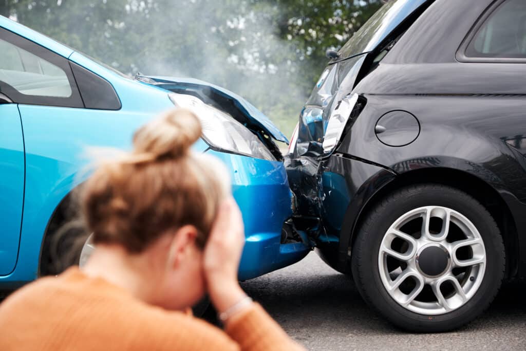 Female Motorist With Head In Hands Sitting Next To Vehicles Involved In Car Accident